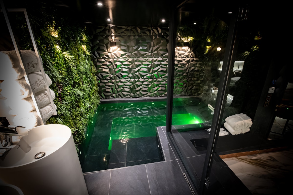 An indoor pool area is showcased, featuring a modern design with a textured dark wall adorned with greenery. Soft green lighting reflects off the water's surface. Neatly stacked towels are visible on the side, complementing the minimalist aesthetic.
