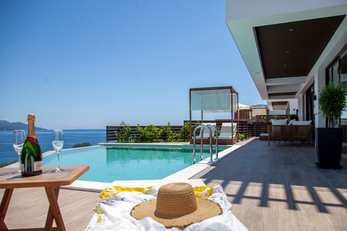 A private pool reflects the clear blue sky, bordered by a spacious terrace with modern seating. A table set with champagne glasses and a straw hat offer a welcoming touch. Lush greenery complements the setting, providing an inviting atmosphere with views of the sea.