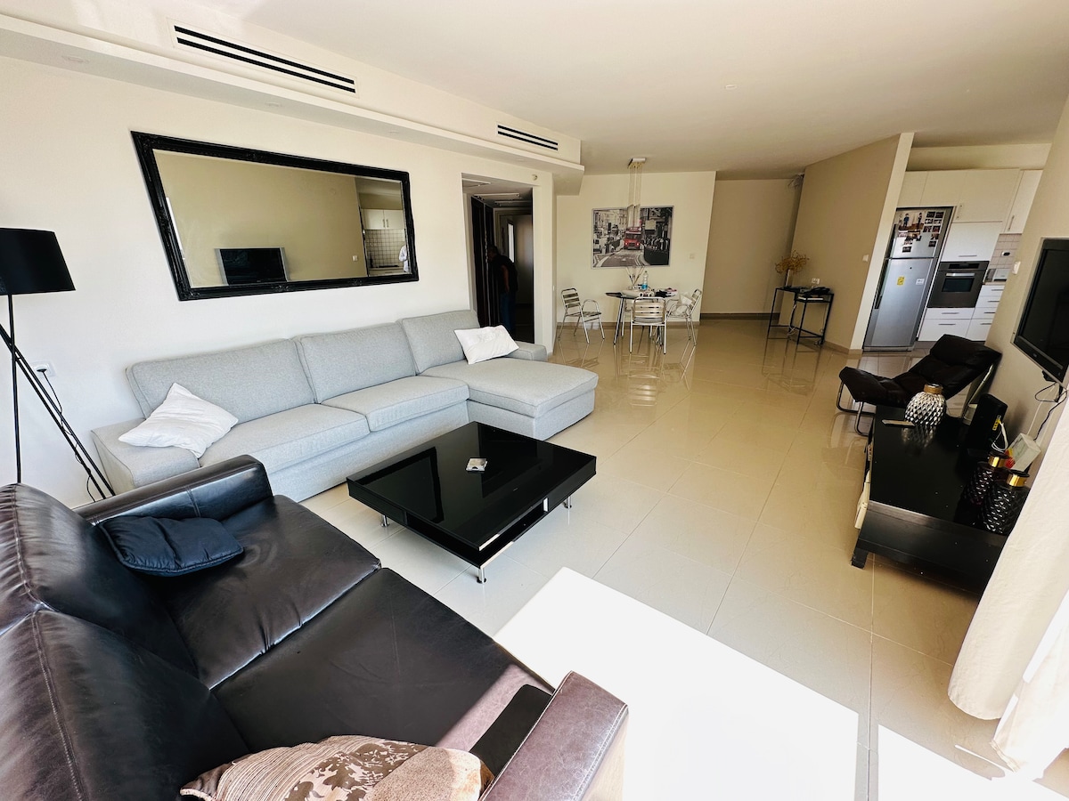 A spacious living area features a light grey sectional sofa facing a dark coffee table. A large mirror reflects natural light, and a dining space with four chairs is visible in the background. Modern appliances are situated in the adjacent kitchen area.