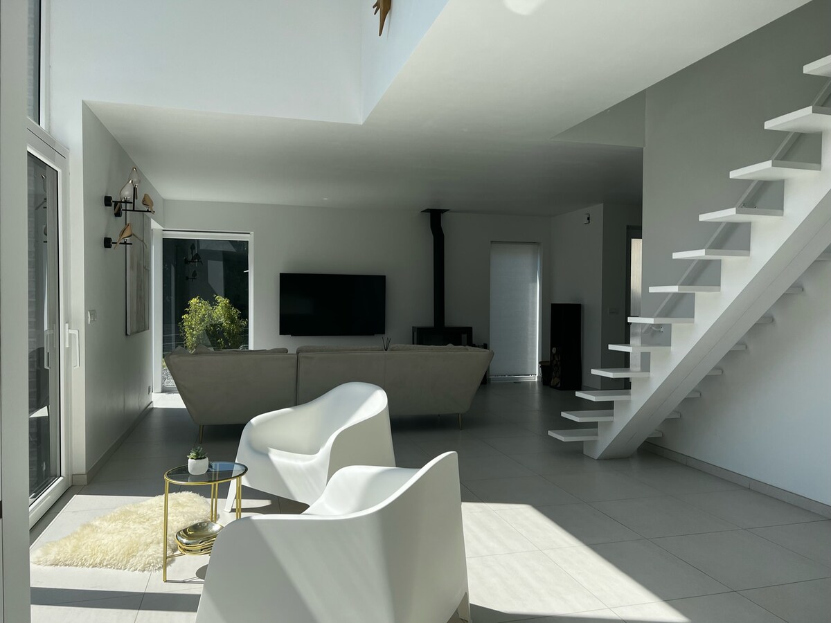 A modern living area is illuminated by natural light through large windows. Two sleek white chairs are positioned beside a small glass table, while a spacious couch faces a minimalist entertainment center. A staircase leads to the upper level, enhancing the open design.
