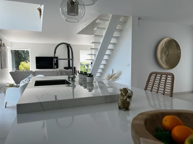 The open-plan living area features a modern kitchen island with a sleek countertop. Natural light floods the space through large windows, highlighting a minimalist design. A staircase leads to the upper level, and a seating area is visible in the background.