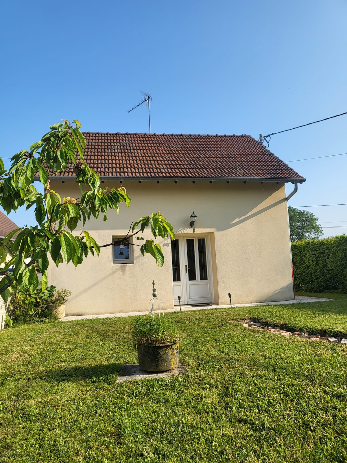 A charming house exterior is featured, displaying a light-colored facade with a sloped roof. A front entryway with double doors is visible, surrounded by well-maintained grass. A small tree and a circular flower bed enhance the simple garden space.