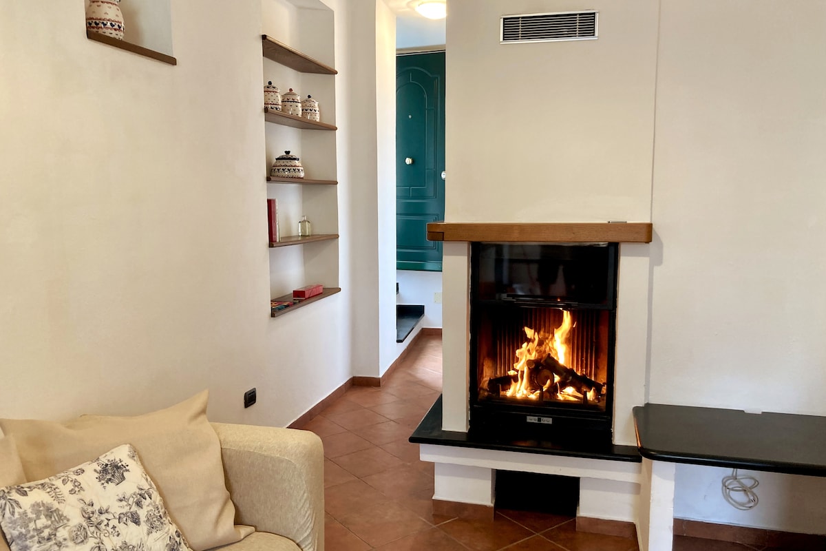 A cozy living area features a traditional fireplace with a warm, flickering flame. A light-colored sofa sits nearby, complemented by a small side table. Decorative shelves display pottery and add character to the neutral walls, while a doorway leads to another area of the home.