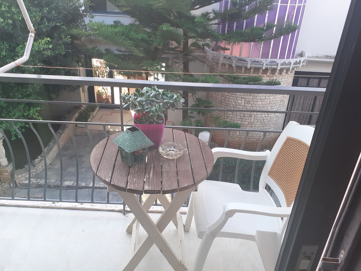A small balcony is featured, showcasing a round wooden table paired with a white chair. A potted plant sits on the table, alongside a decorative item. Lush greenery is visible in the background, creating a serene environment.