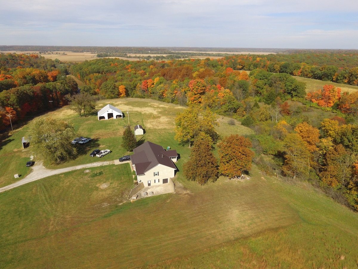 An aerial view captures a spacious property surrounded by vibrant autumn foliage. The home, positioned prominently, is complemented by a large outbuilding and ample open space. The landscape features gentle rolling hills under a clear sky, creating a peaceful natural setting.