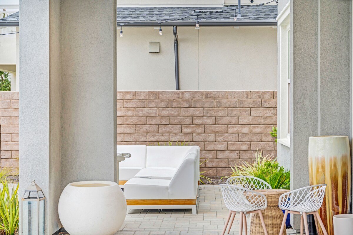 An outdoor seating area features a white sectional sofa, surrounded by decorative planters and textured walls. The paved stones create a neat pathway, while the space is enhanced by soft lighting fixtures above, inviting a relaxed atmosphere for social gatherings.