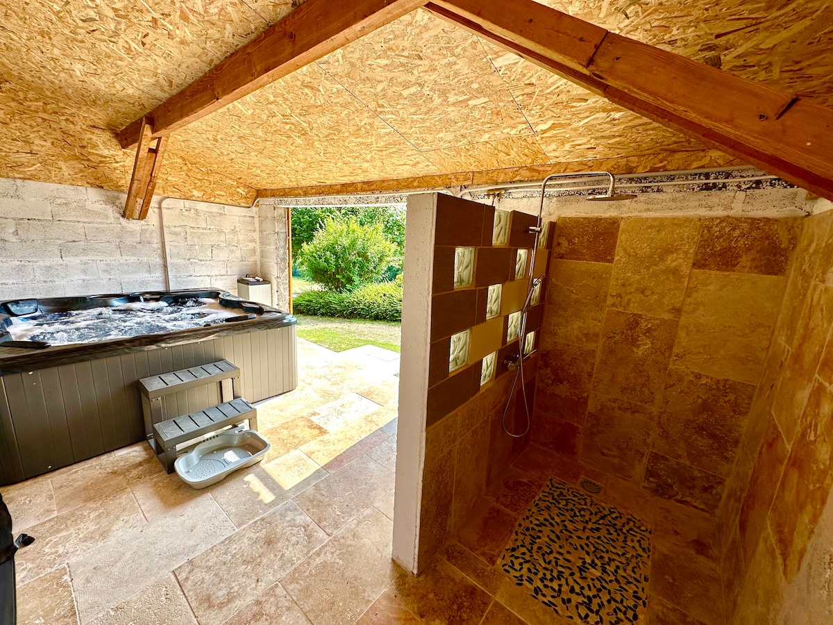 An outdoor wellness area features a hot tub adjacent to a tiled shower space. Natural light enters through the open doorway, highlighting the earthy tones of the stone tiles. The structure is accented by an overhead wooden beam and a textured ceiling.