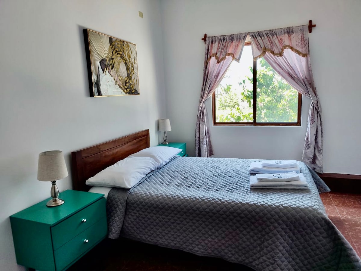 A well-lit bedroom features a spacious bed with white bedding, flanked by two turquoise nightstands. Soft curtains frame a window that allows natural light to enter, illuminating the room. A piece of artwork hangs above the bed, adding a touch of character.