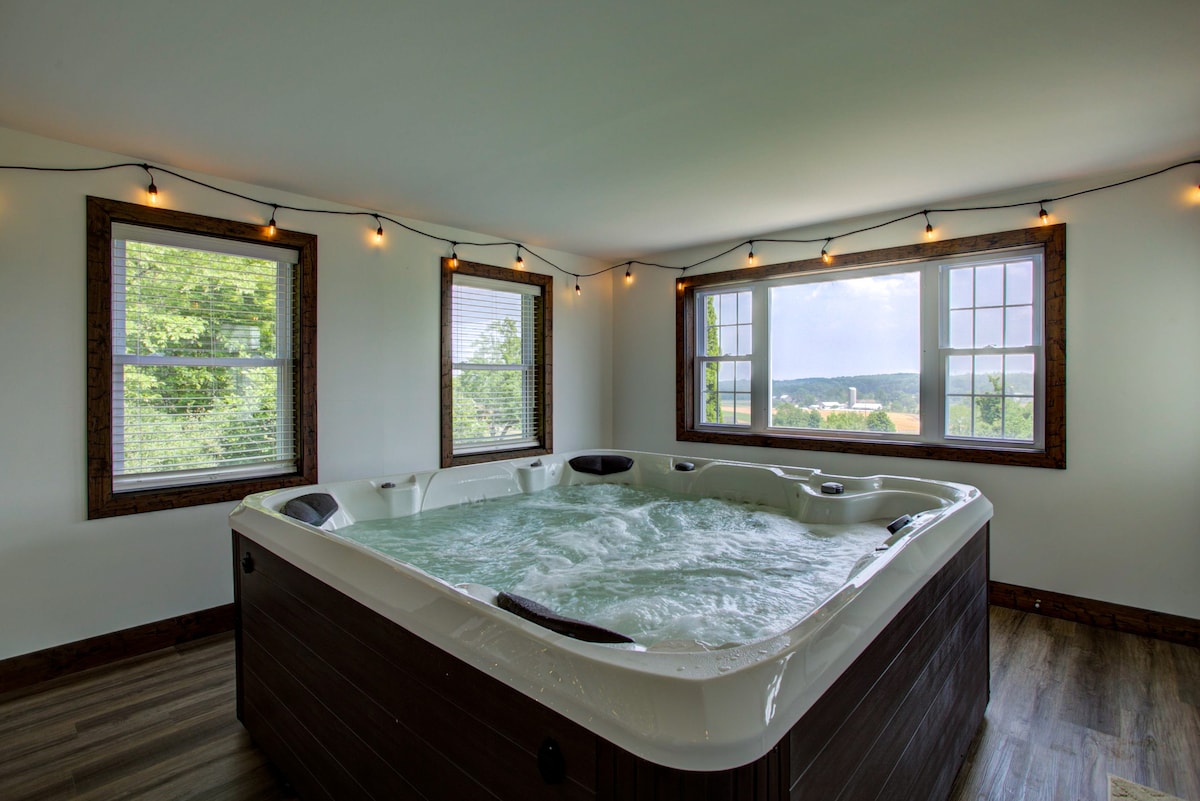 A spacious hot tub is positioned in front of large windows, providing expansive views of the surrounding landscape. Soft string lights hang from the ceiling, enhancing the relaxed ambiance in the well-lit space.