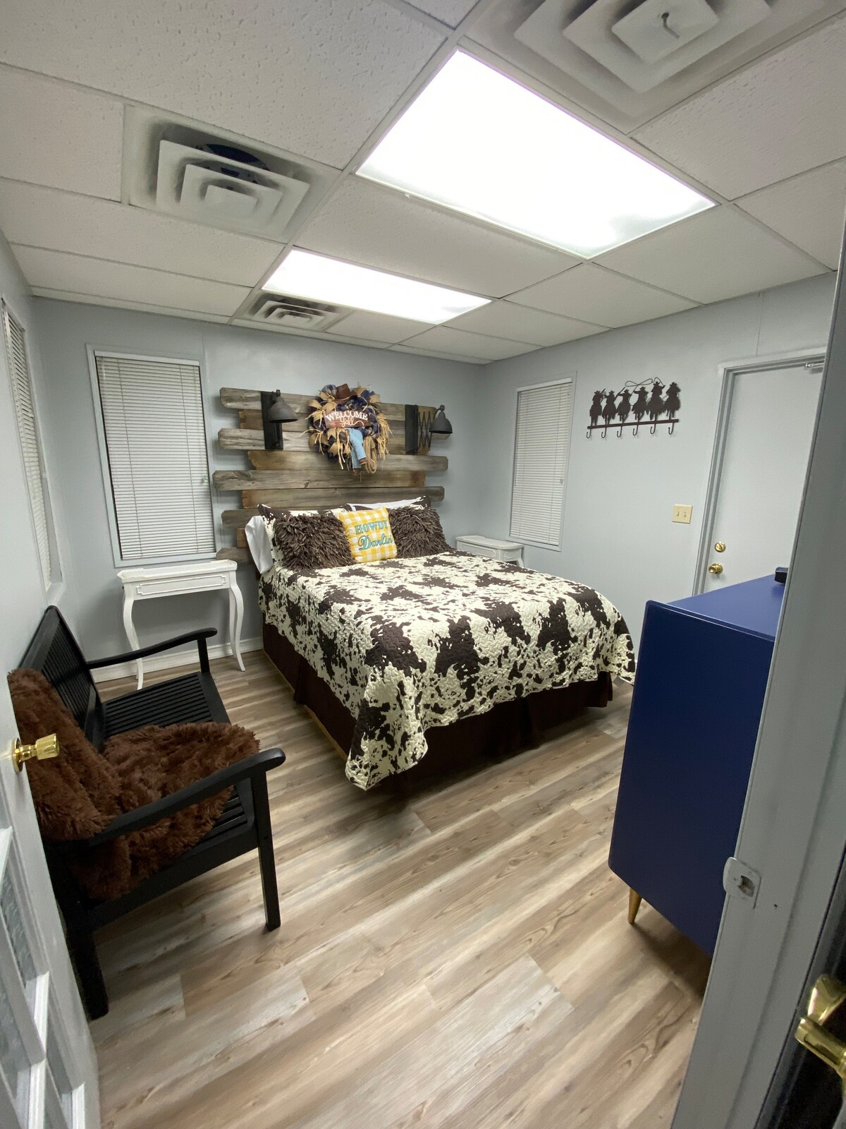 A well-organized bedroom features a queen bed with a patterned quilt and a charming wooden headboard. A cozy bench is placed against the wall, and a small table adds functionality. Natural light floods in through the window, complemented by minimalist decor.