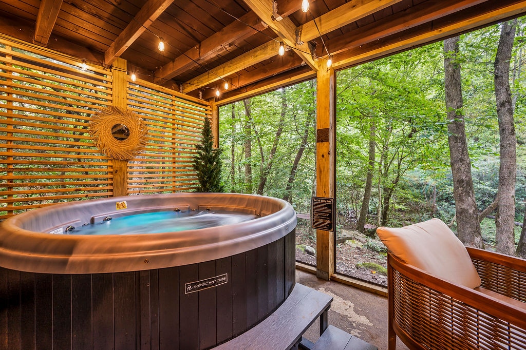 A hot tub is centrally located on a wooden deck, surrounded by greenery. Soft lighting highlights the space, while large windows provide views of the forest. Natural wood elements are featured throughout, creating a serene atmosphere for relaxation.
