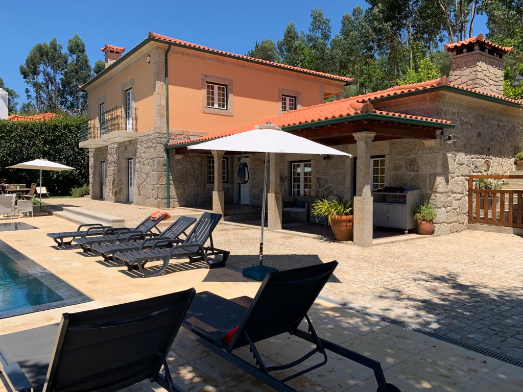 A charming house constructed with stone and adorned with vibrant orange accents is situated beside a serene pool. Loungers surround the pool area, and an umbrella provides shade. The outdoor space is bordered by greenery, enhancing the peaceful atmosphere.