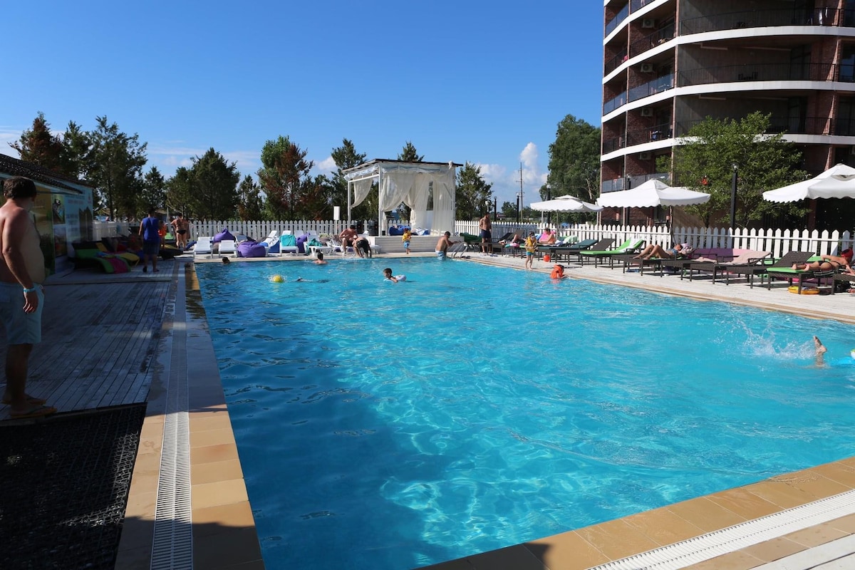 A spacious outdoor pool is surrounded by lounge chairs and umbrellas. Several guests are seen enjoying the water, while others relax alongside the pool. Lush greenery and a modern building are visible in the background, contributing to the vibrant atmosphere.