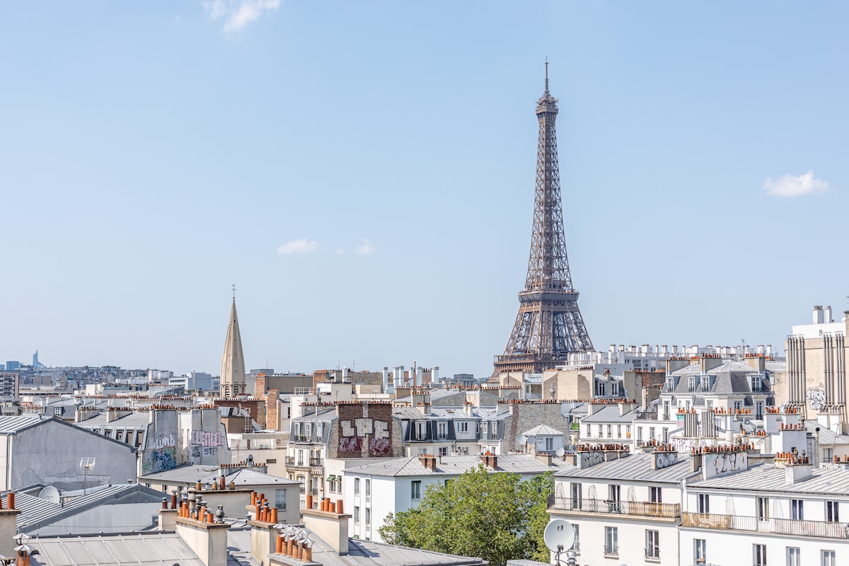 A panoramic view of the iconic Eiffel Tower rises above a landscape of Parisian rooftops. Various building styles are visible, featuring characteristic French architecture, with chimneys adorning the roofs. The sky is clear, enhancing the cityscape experience.