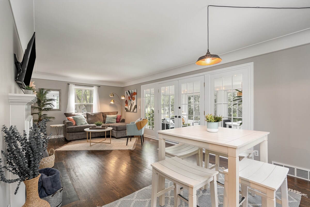 The living area features a comfortable gray sectional sofa and a flat-screen television mounted on the wall. A wooden dining table with three chairs is positioned nearby. Natural light enters through multiple windows, enhancing the open and airy feel of the space.