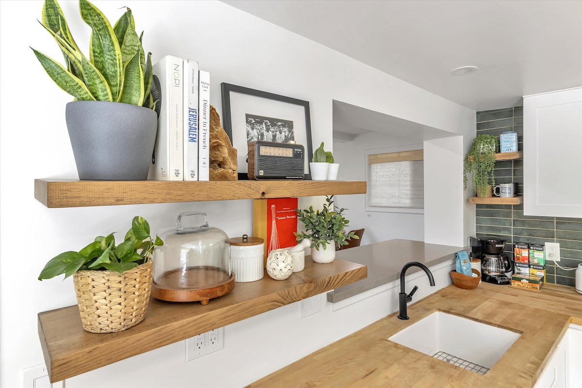 A sleek kitchen area is highlighted by wooden shelving displaying plants and books. Below, a sink is positioned against a backdrop of modern appliances, while a coffee station is visible on one side, creating an organized and functional space.