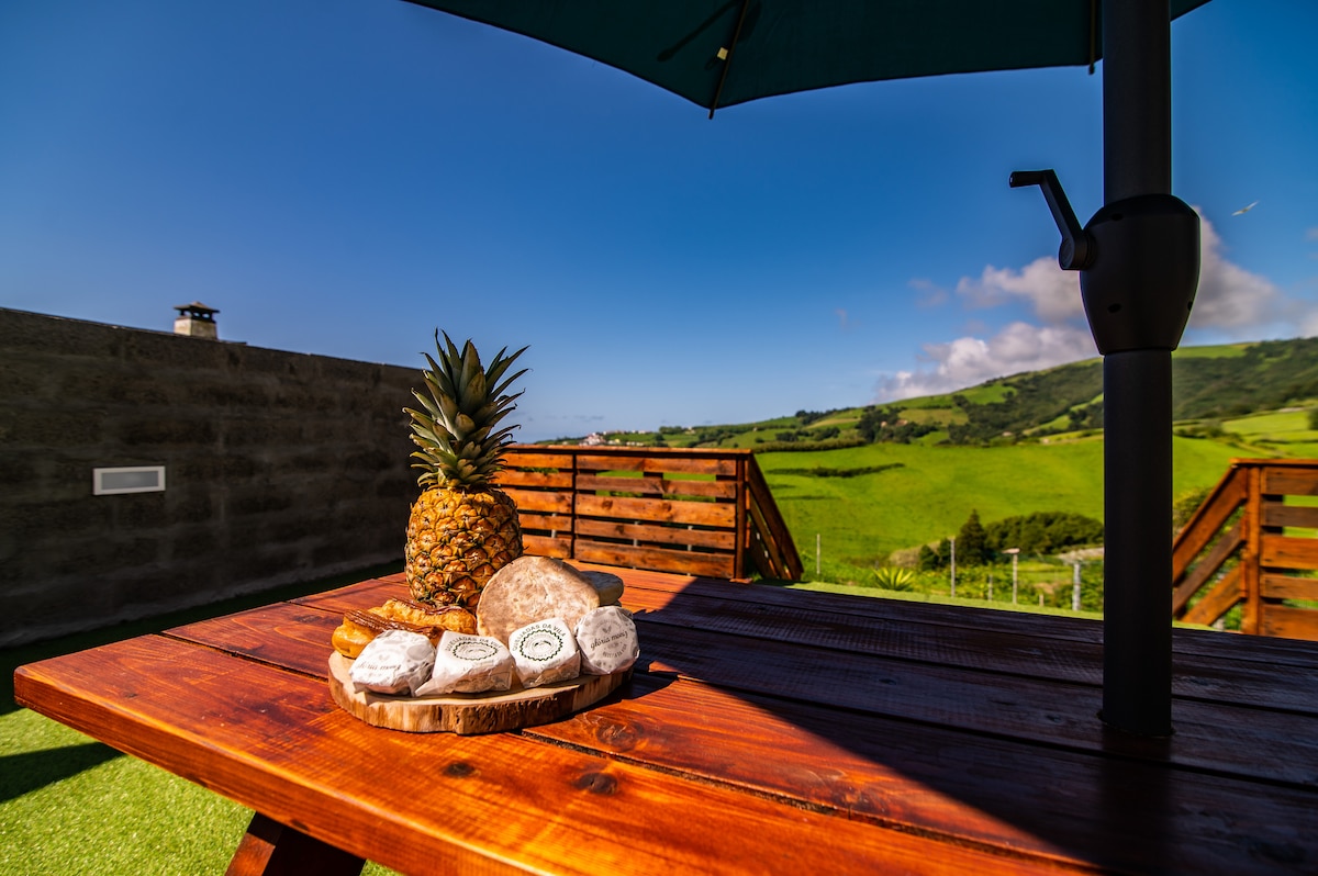 A wooden table is set under a shaded umbrella, featuring a selection of local cheeses and a pineapple centerpiece. Lush green hills extend in the background, complemented by a clear blue sky.