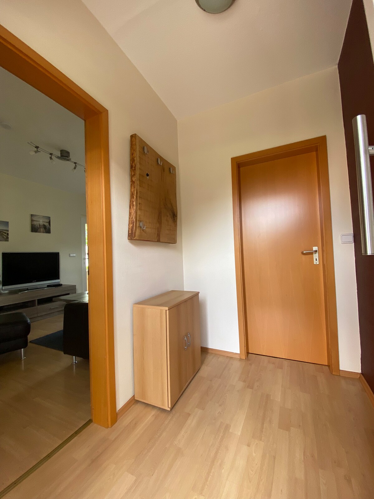 A bright hallway features wooden accents and light-colored walls. A wooden cabinet is positioned to one side, providing storage space. A door leads to another room, while a mirror reflects the scene. Natural light enters through a nearby window.