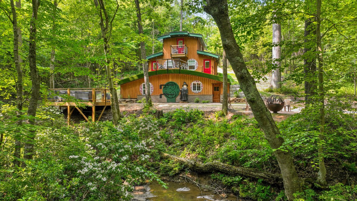 The Hobbit House, designed with a distinctive architecture, is nestled among lush greenery. The wooden structure features multiple levels and vibrant accents, with windows and a deck that integrate seamlessly with the natural surroundings. A small stream flows nearby, enhancing the tranquil setting.