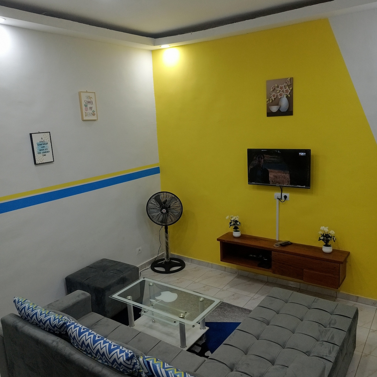 A modern living room features a gray sectional sofa facing a wooden entertainment unit with a television. A glass coffee table sits in front, while a standing fan provides ventilation. The walls are painted in yellow and white, accented by blue stripes and framed artwork.
