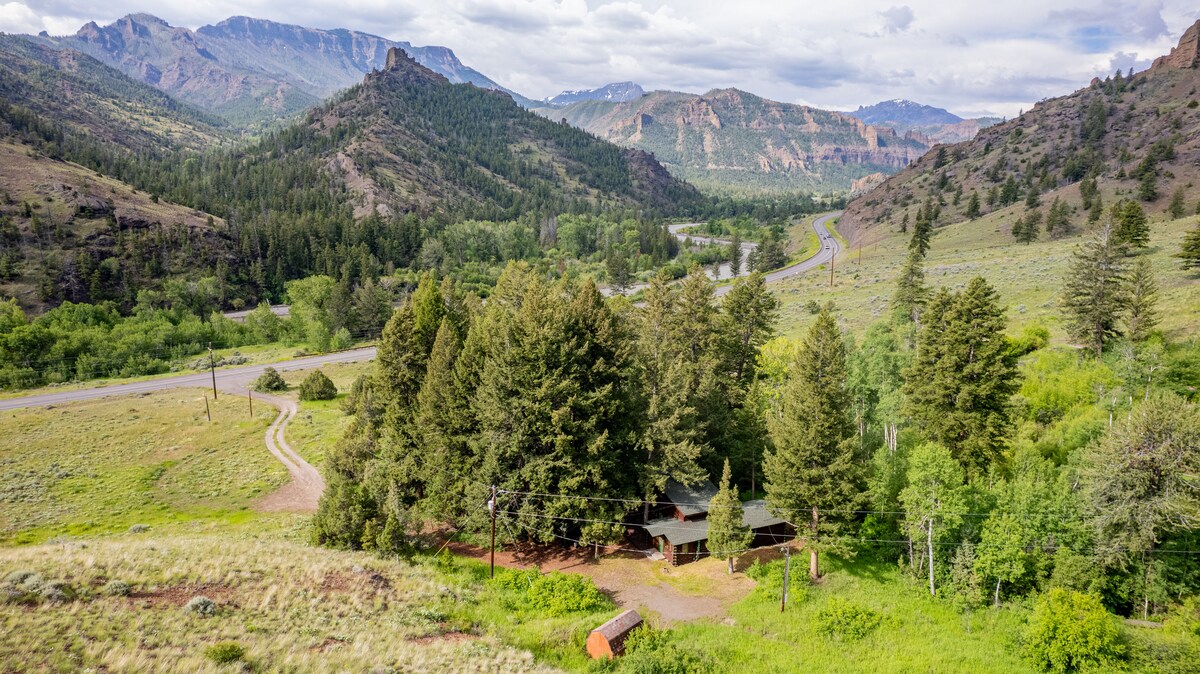 A secluded cabin is nestled within lush greenery, surrounded by tall trees and mountainous terrain. A winding road can be seen nearby, leading into the forested landscape, while the North Fork of the Shoshone River flows gently along the valley below.