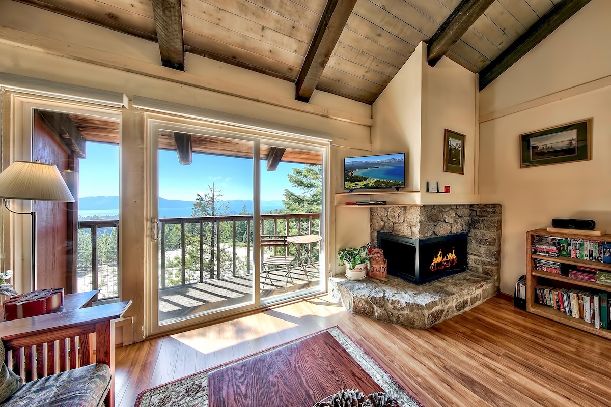 A cozy living area features a stone fireplace and large sliding glass doors offering expansive views of the lake and surrounding trees. A television is mounted on the wall, and a small table and chairs are positioned on the balcony. Natural light illuminates the wooden beams and floors.