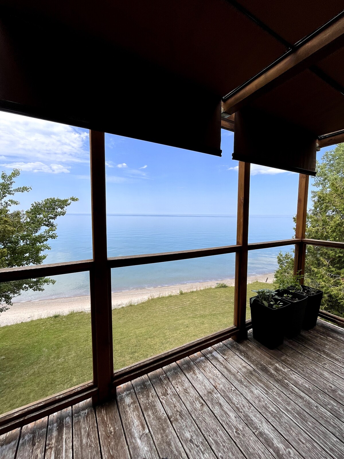 A serene view is displayed from a screened porch, featuring large windows that overlook Lake Huron. The gently sloping beach and calm water are visible, surrounded by greenery, offering an inviting outdoor space. Potted plants are positioned along the wooden deck, blending with the natural setting.
