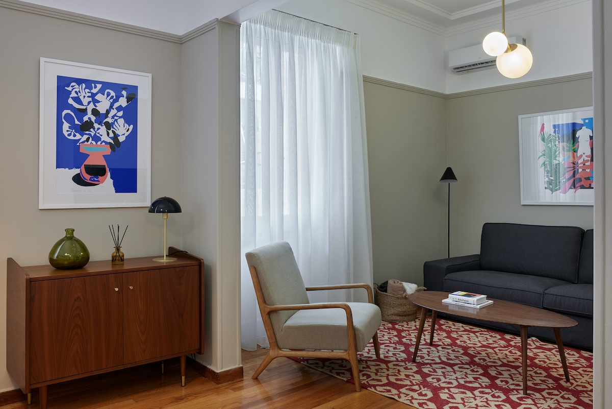 A thoughtfully arranged living area is presented with a dark sofa and a light armchair positioned around a low wooden table. Natural light enters through a large window dressed in sheer curtains. Decorative wall art adds a touch of color to the neutral-toned walls.