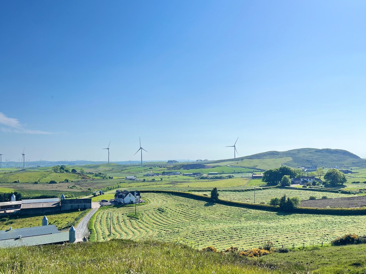 A panoramic view of rolling green fields is presented, with several wind turbines in the distance. A small white house is visible along with scattered trees, and a gentle hill rises in the background under a clear blue sky.