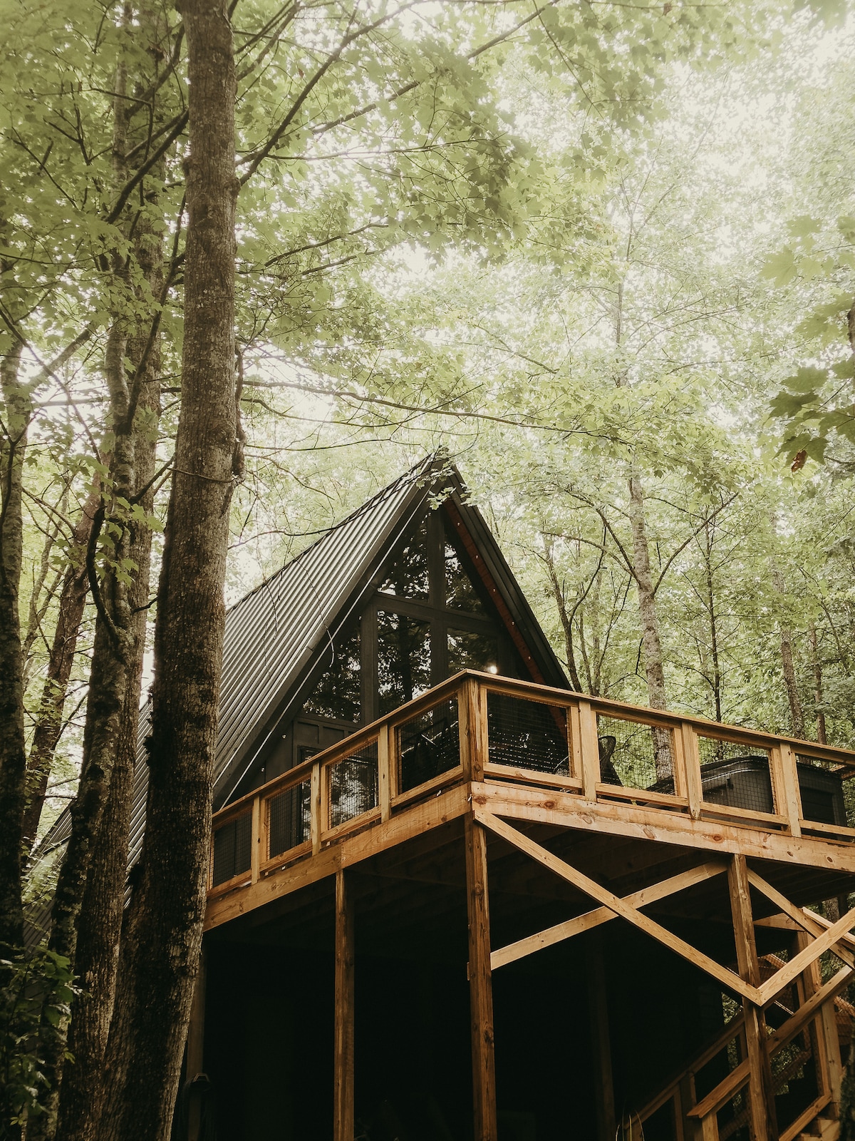 The exterior of an A-frame cabin is showcased amid lush green trees. The structure features a wooden deck with railings, blending harmoniously with the surrounding forest. Natural light filters through the canopy above, creating a serene atmosphere.