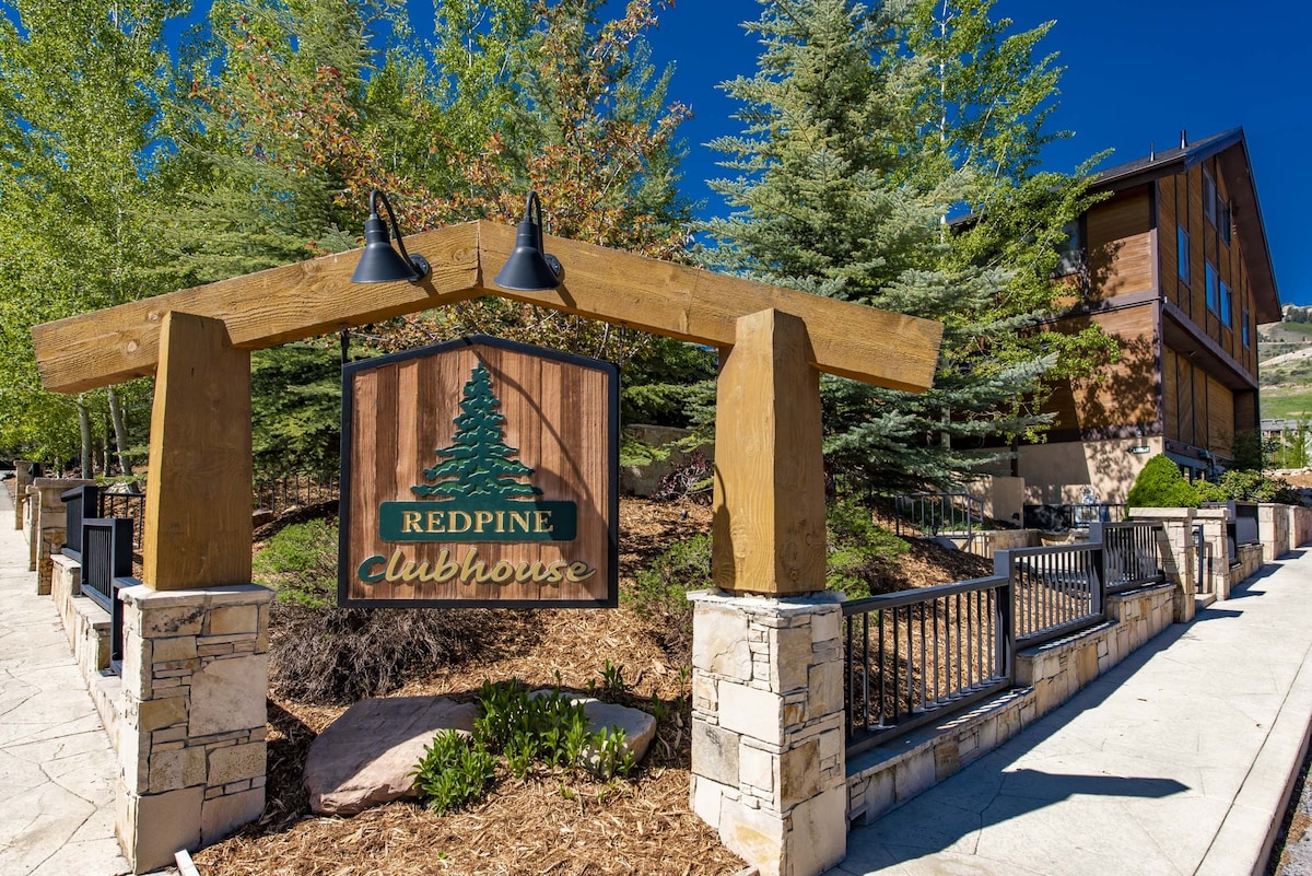 The entrance to the Red Pine Clubhouse is marked by a wooden sign featuring a tree logo. Surrounding greenery and landscaped rocks create a natural setting. Two light fixtures hang above the sign, while the building’s rustic architecture offers a welcoming presence.