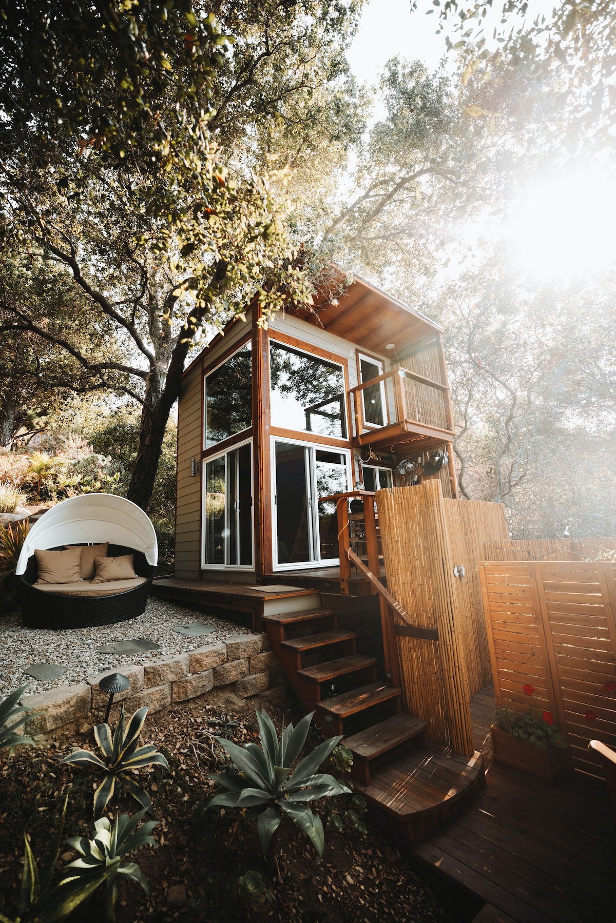 A charming tiny house is nestled among trees, featuring large glass windows that provide a bright and airy feel. A wooden ladder leads to a balcony, surrounded by greenery. A cozy outdoor seating area with a round, cushioned chair complements the natural setting.