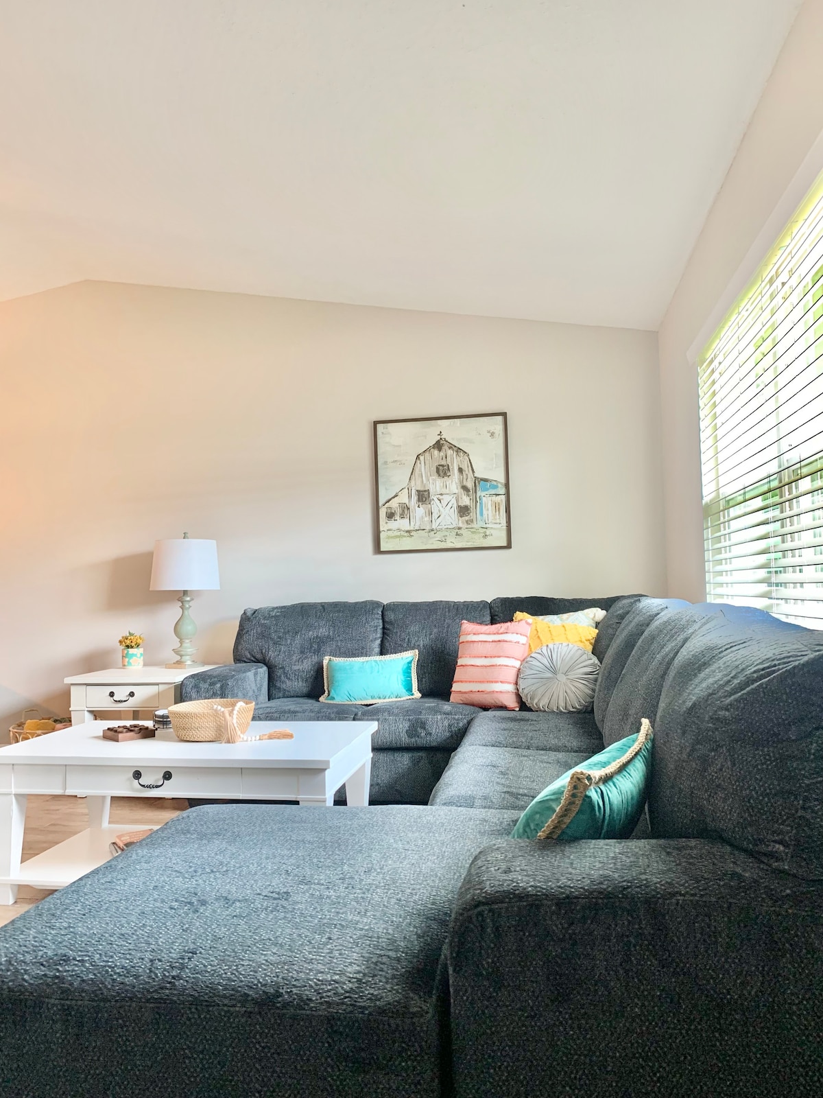 A spacious living area is furnished with a large sectional sofa, accented by colorful throw pillows. A white coffee table stands in front, accompanied by a decorative basket. Natural light filters in through the nearby window with white blinds.