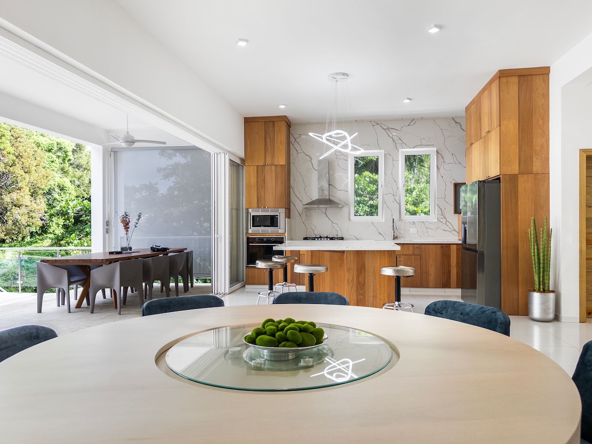 An open-concept kitchen and dining area showcases modern wooden cabinetry paired with sleek marble accents. A round dining table is centered in the room, complemented by blue chairs. Large windows bring in natural light and offer views of the surrounding greenery.