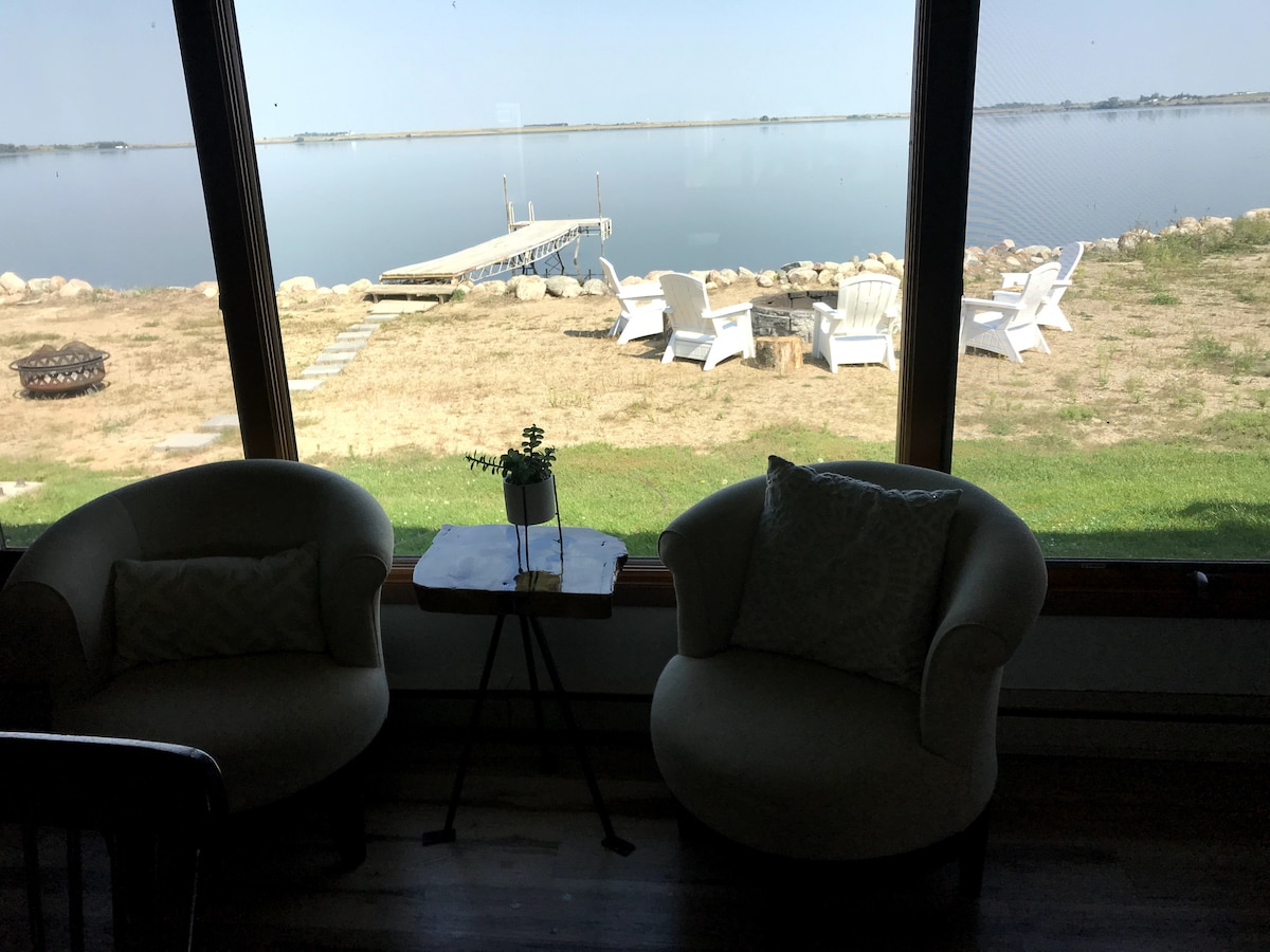 A serene corner of the living space is highlighted by two comfortable chairs positioned near a large window. Outside, a private dock extends into the calm waters of Bluedog Lake. Several white outdoor chairs are visible along the shoreline, creating a peaceful lakeside setting.