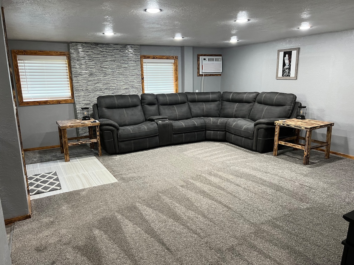 A spacious living room features a large L-shaped sectional couch in a dark color, complemented by two rustic wooden side tables. The room is well-lit with recessed lighting and natural light from the windows, and a neutral carpet covers the floor.