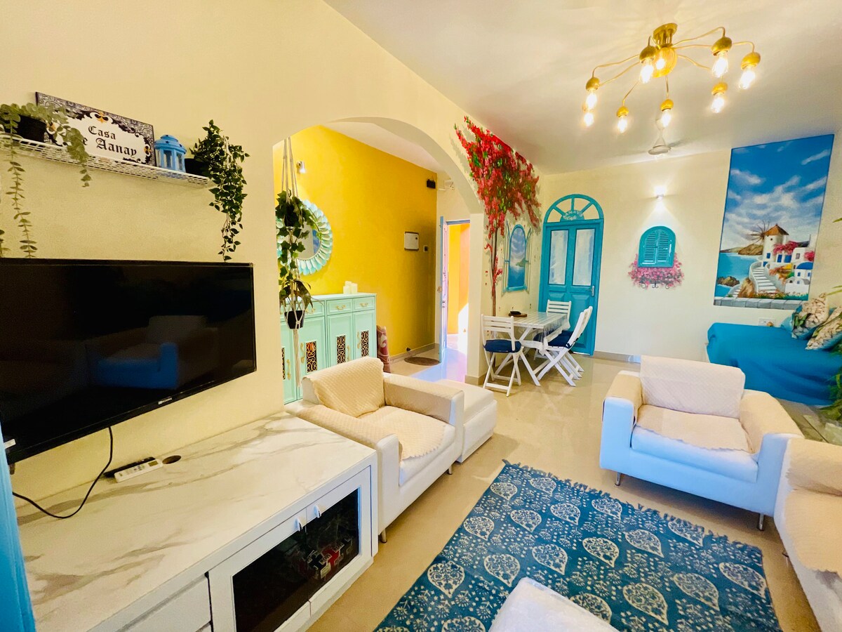 A bright living area features a large TV mounted on a white cabinet, surrounded by stylish seating. Colorful decor includes hand-painted murals and potted plants, while a dining table and chairs are visible in the background, enhancing the open-concept layout.