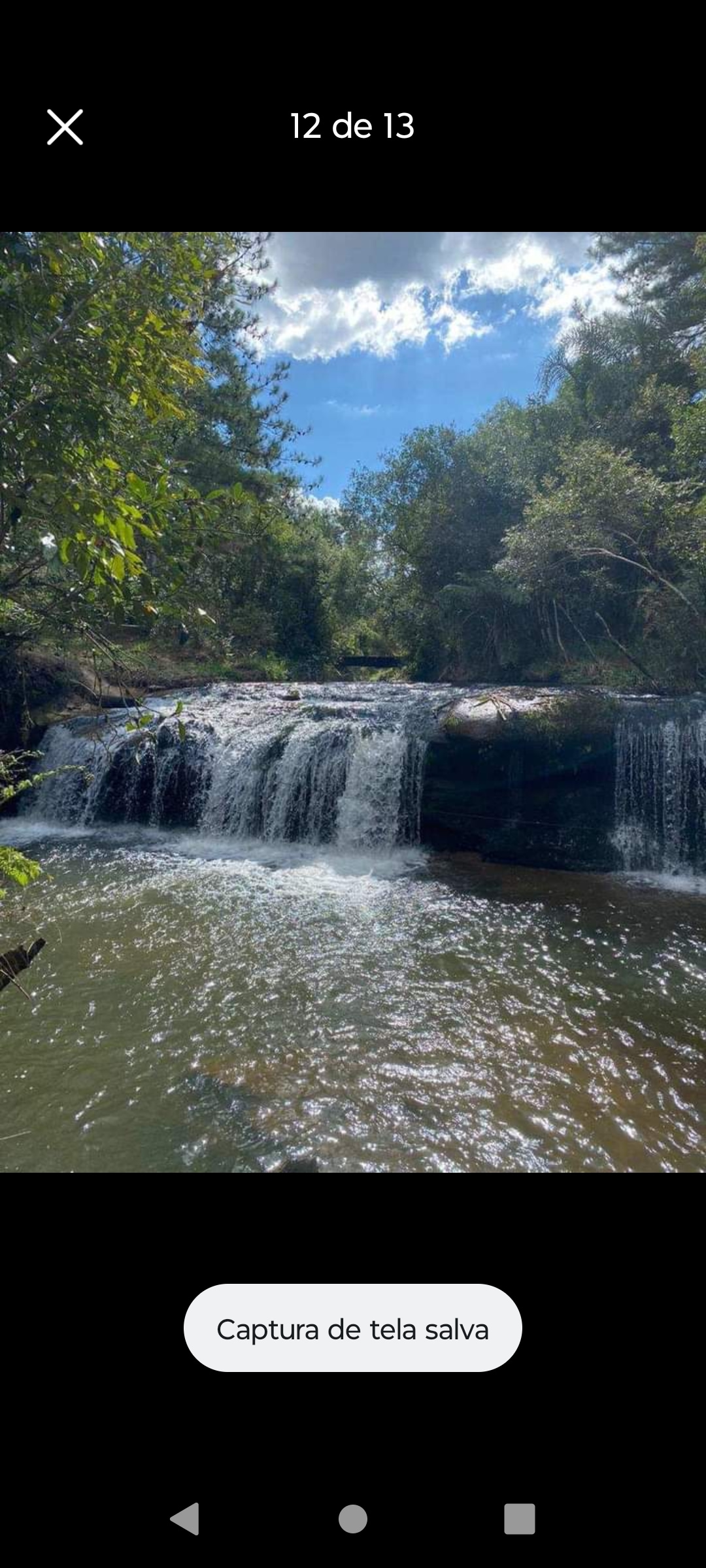 A cascading waterfall is seen flowing over rocky ledges, creating a tranquil ambiance with splashes of water. Lush greenery frames the scene, with trees and foliage surrounding the water's edge, reflecting in the calm river below under a clear blue sky.