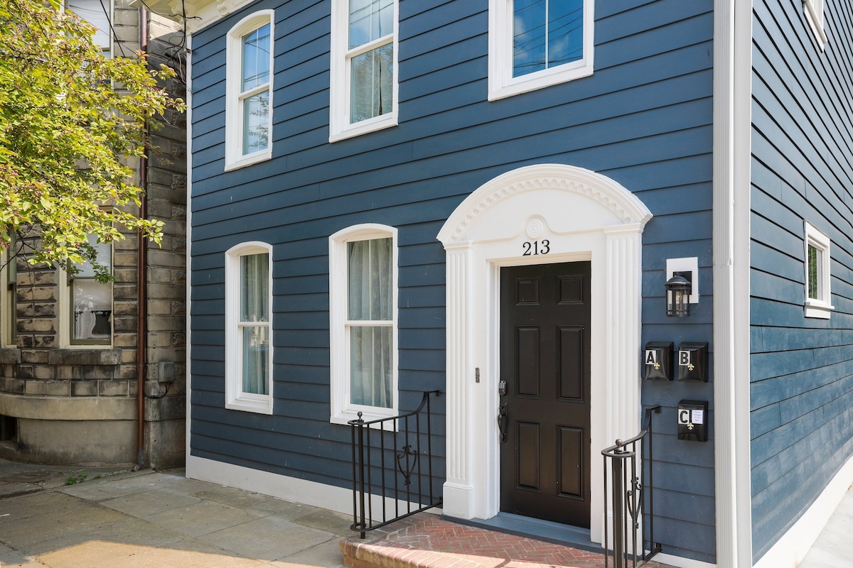 The exterior of the historic apartment is displayed, featuring a bold blue facade and white trim. The front entrance is framed with ornate details, and a black door with a modern handle is highlighted. There are large windows allowing natural light to enter.