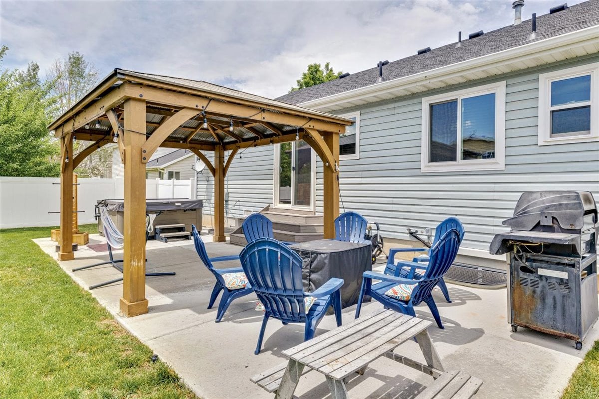 An outdoor patio area is enhanced by a wooden pergola, offering shade over seating arrangements with colorful chairs. A hot tub is positioned nearby, alongside a BBQ grill and picnic table, providing a space for relaxation and dining in a landscaped backyard.