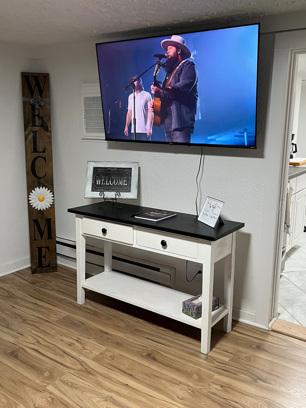 A wall-mounted television displays a live performance, positioned above a white console table with two drawers. The table is placed on a wood floor, complemented by a large 'WELCOME' sign and framed artwork nearby. Air conditioning is visible, enhancing comfort in the space.