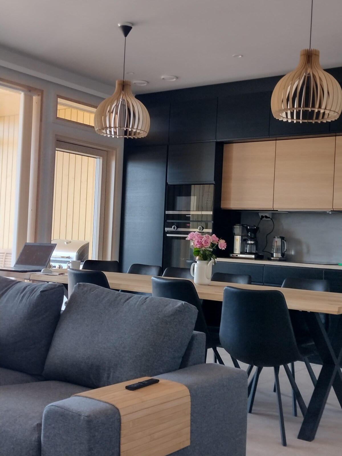 A modern kitchen and dining area is shown, featuring dark cabinetry complemented by light wood accents. A spacious dining table is surrounded by black chairs, and pendant lights hang overhead. A comfortable sofa is positioned nearby, with a decorative flower arrangement on the table.