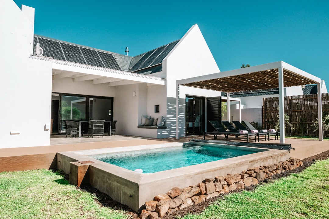A modern villa exterior showcases a crystal clear swimming pool, surrounded by a wooden deck and a low stone wall. A shaded lounge area with sun loungers provides a relaxing space. The roof features solar panels, complemented by large glass doors that open into the interior.