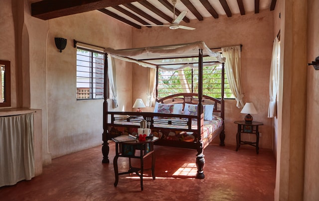 A spacious bedroom features a four-poster bed draped with light fabric. Two bedside tables with lamps are positioned beside the bed, and windows allow natural light to fill the room while providing views of the greenery outside.