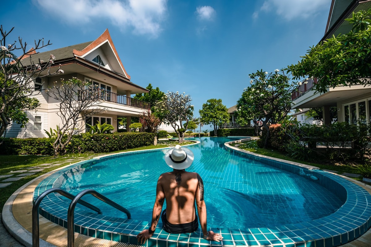 A serene salt-water pool is surrounded by lush greenery and elegant homes. The pool's inviting turquoise water reflects the blue sky above, creating a tranquil setting. Sunlight highlights the gentle curves of the pool's design, while shaded areas provide comfort for relaxation.