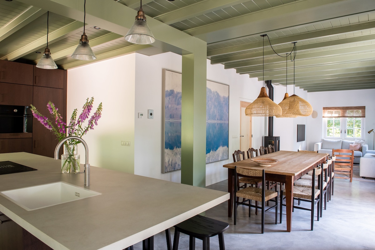 An open-plan living area features a large wooden dining table surrounded by chairs, with pendant lighting overhead. An island kitchen with a sink highlights a minimalist design. Natural light enters through windows, illuminating the space and enhancing the green ceiling beams.
