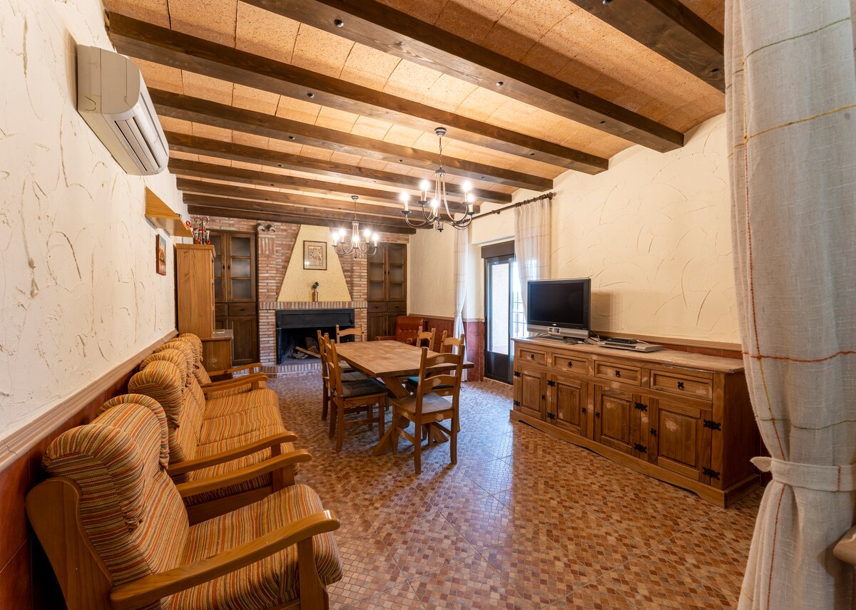 A spacious living area features wooden beams across the ceiling, with a large dining table surrounded by chairs. Two comfortable seating areas include armchairs and a sofa. A rustic wooden cabinet and a television are visible, enhancing the cozy ambiance of the room.