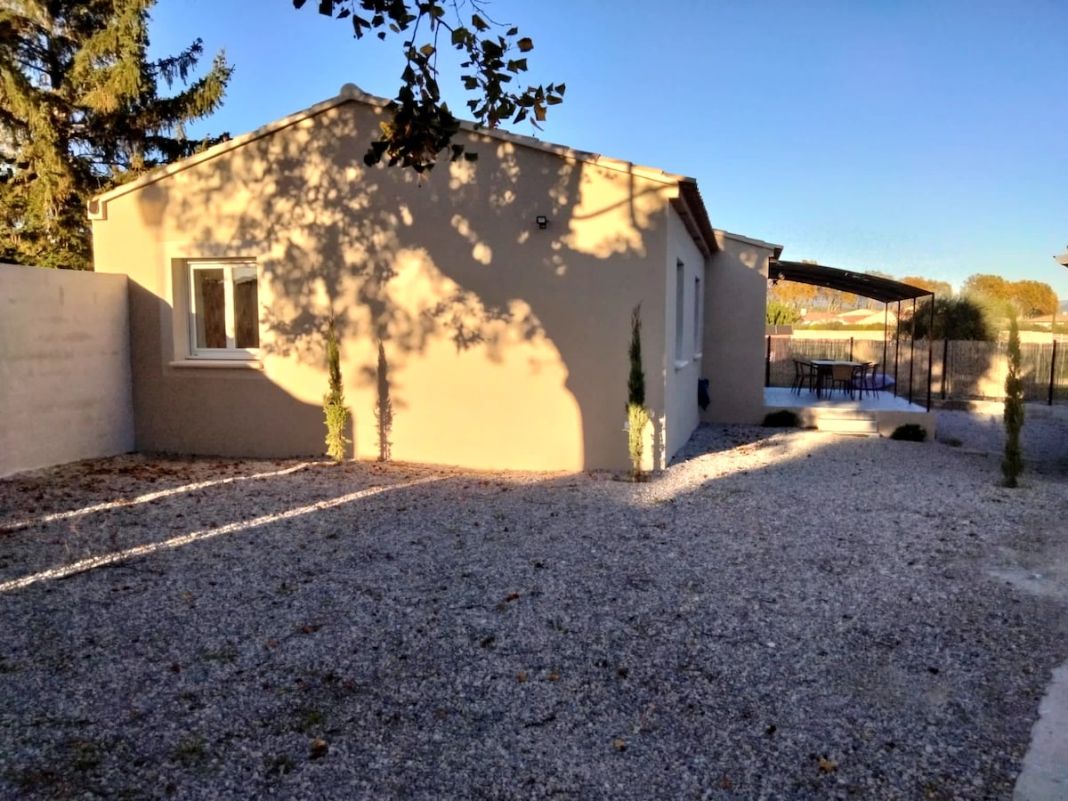 The exterior of a newly constructed villa is shown, featuring a light-colored façade and a gravel yard. Tall trees and small shrubs are placed along the perimeter, enhancing the landscape. A shaded terrace area is visible, offering outdoor seating options.