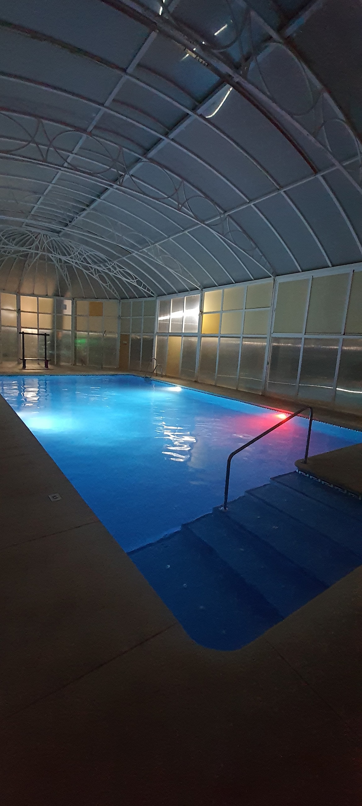 An indoor heated pool is featured, surrounded by glass panels that allow natural light. The water is illuminated with a soft blue hue, enhancing the calm atmosphere. Stairs lead into the pool, providing easy access.
