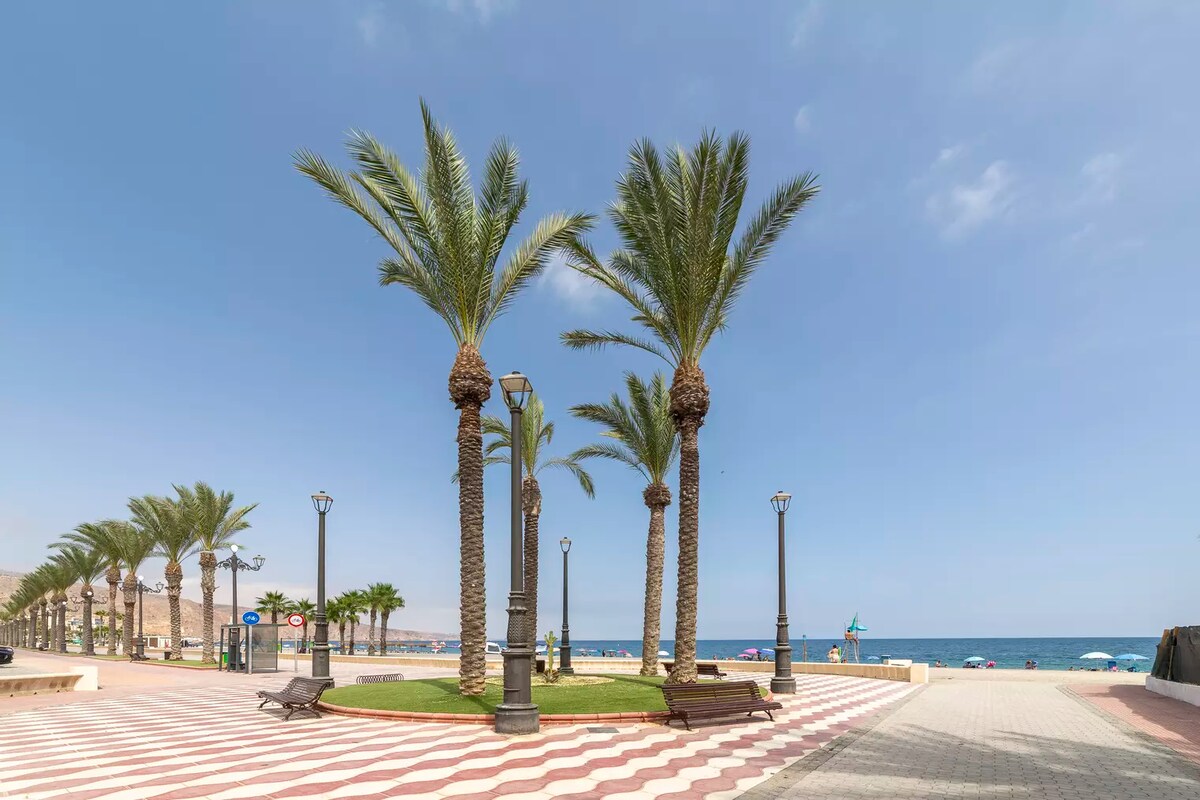 A beachfront promenade is lined with palm trees, offering shade along the walkway. The sea can be seen in the distance, with umbrellas dotting the sandy shore. The ground features a patterned surface, enhancing the outdoor space for leisurely strolls.